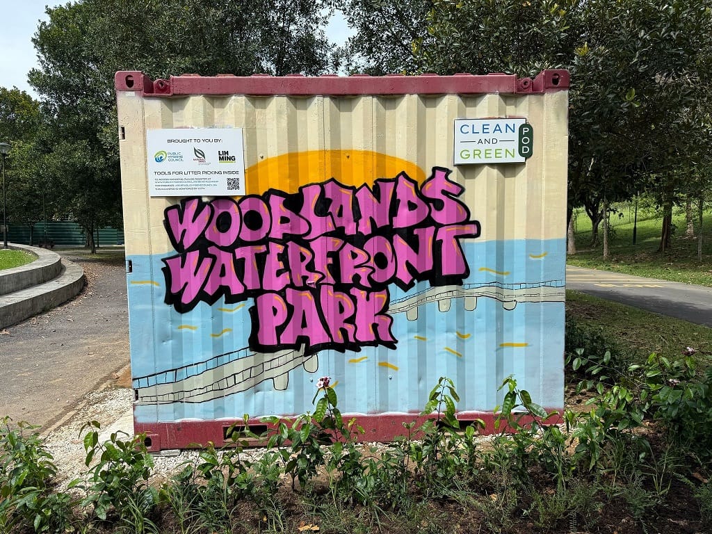 A CleanPod container at Woodlands Waterfront Park, decorated with a colourful graffiti-style mural featuring the words 'Woodlands Waterfront Park' in pink lettering, with a scenic waterfront and bridge illustration in the background. A 'Clean and Green' label and a notice indicating litter-picking tools are available inside can be seen on the container.